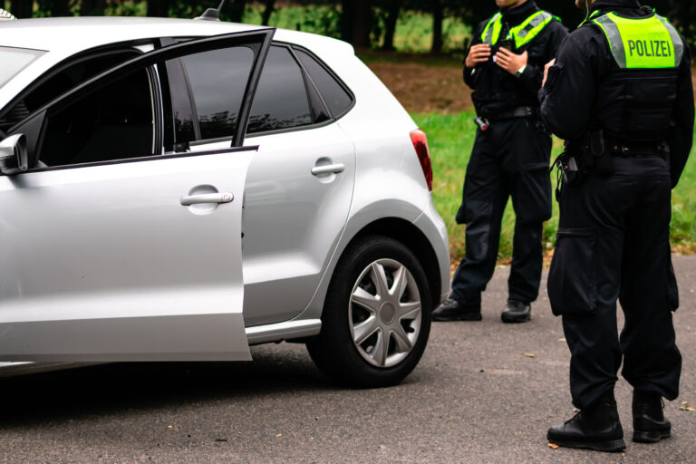 Side view of a silver car with the rear door open and two police officers in black uniforms with neon vests labeled POLIZEI nearby.
